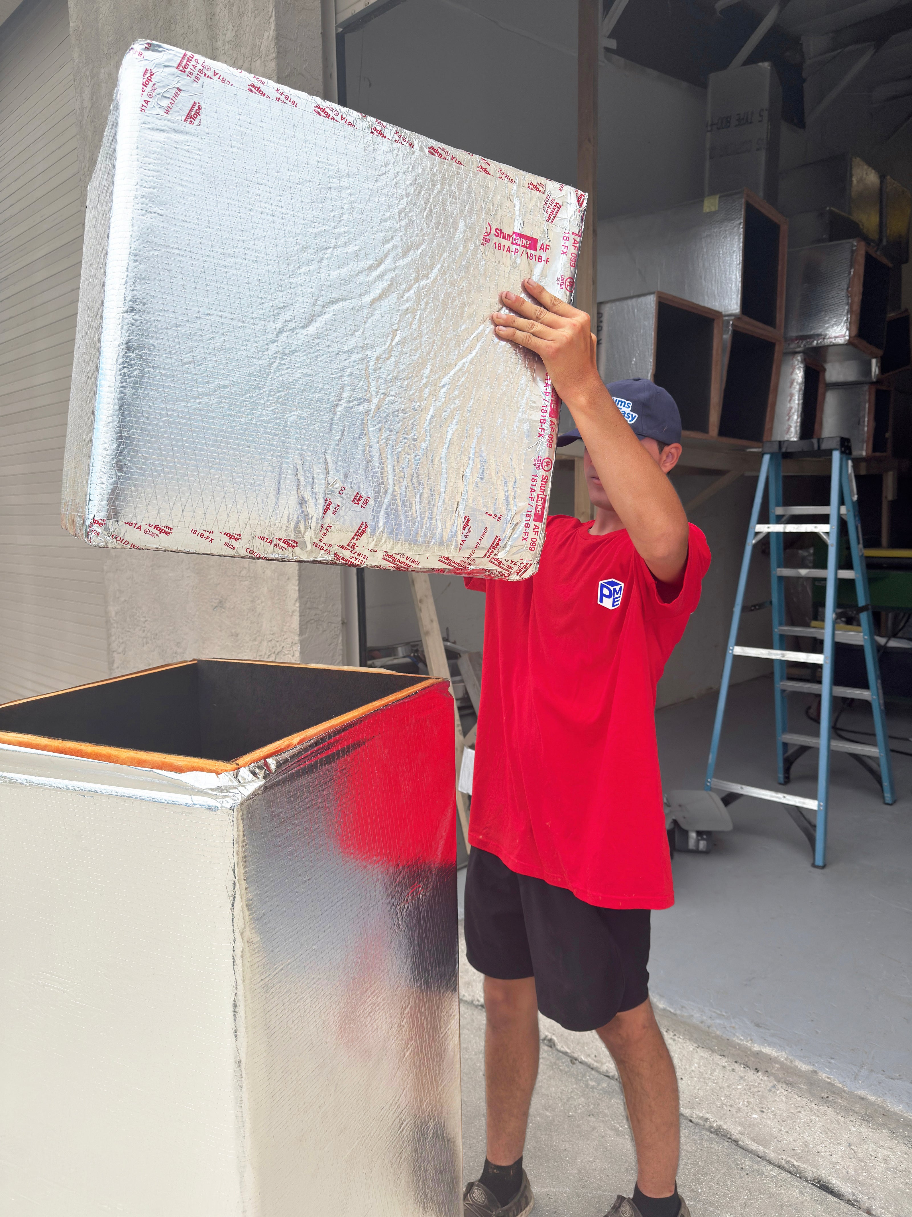 Person in a red 'Plenums Made Easy' shirt lifting a large duct board plenum in an industrial setting